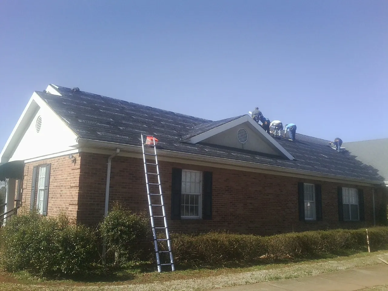 Crew tearing shingles off a brick ranch roof with a ladder set — Emergency Roof Repair work by our crew in Belmont
